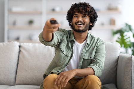 Cheerful indian guy sitting on couch, watching TV