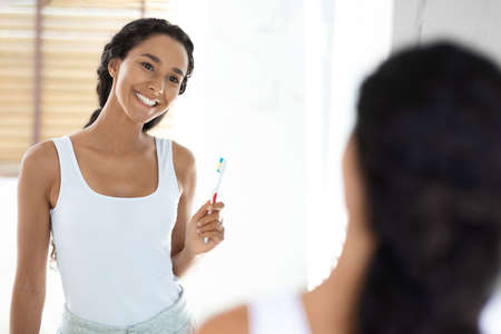 Happy Young Female Holding Toothbrush And Looking At Mirror In Bathroom