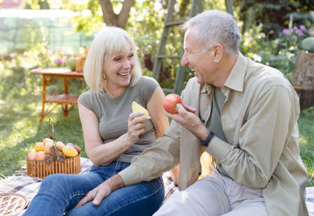 Laughing senior spouses sitting on field grass and eating fruits, talking and smiling, resting...
