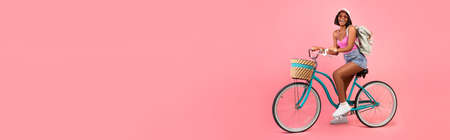 Black woman in casual summer clothes riding bicycle over pinkの写真素材