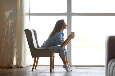 Side view of beautiful young woman drinking hot coffee, sitting on chair near window at home, free spaceの写真素材