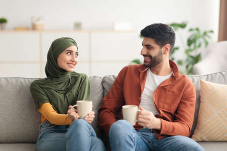 Happy Arabic Spouses Holding Mugs Drinking Coffee Sitting At Homeの写真素材
