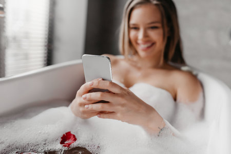 Cheerful woman using smartphone while lying in foamy bath, reading beauty blog or watching video online and smilingの写真素材