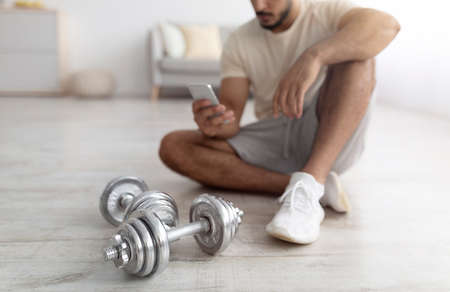 Unrecognizable young Arab man using mobile phone on break from domestic strength training, selective focus on dumbbellsの写真素材