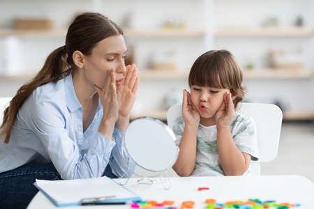 Cute little boy with speech problems practicing with therapist, doing practice and looking at...