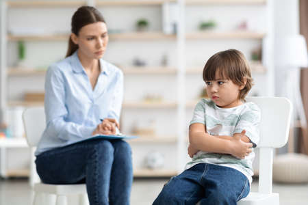 Offended little boy sitting with folded arms, neglecting communication with woman psychologist during consultationの写真素材