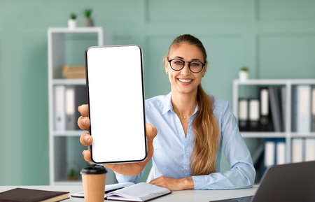 Positive businesswoman showing modern cellphone with big blank screen, sitting at workplace in office, mockupの写真素材