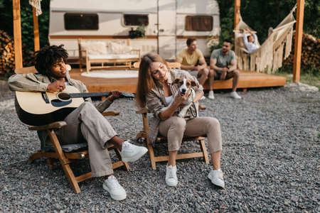 Young multiethnic people relaxing in lounge chairs with guitar and cute pet dog, having fun vacation near RV, copy spaceの写真素材
