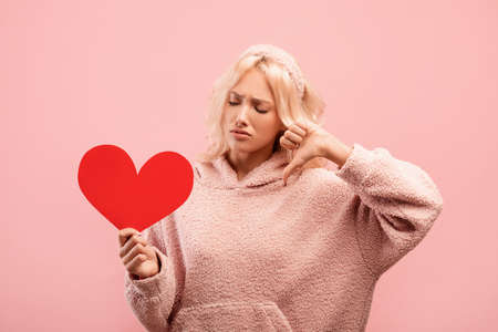 Sad and heart broken young lady holding red paper heart and showing thumb down, posing over pink...