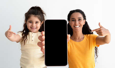 Arab Mom And Cute Little Daughter Pointing At Smartphone With Black Screen