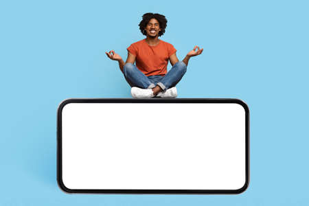 Peaceful black guy meditating, sitting on big cell phone, mockup