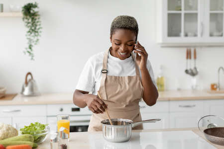 Happy millennial african american woman in apron prepare food and making phone call in modern scandinavian kitchenの写真素材