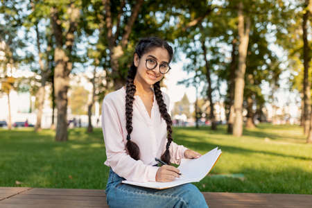 Happy indian millennial girl sketching or taking notes, doing homework while sitting in park and smiling at cameraの写真素材