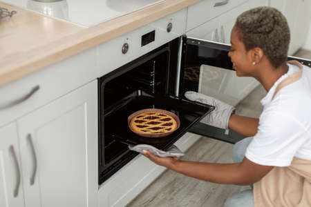 Happy millennial african american female in apron takes out pie from oven in minimalist kitchen...