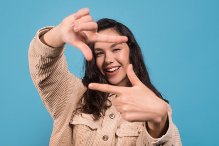 Cheerful young pretty european lady looking through fingers like camera isolated on blue backgroundの写真素材