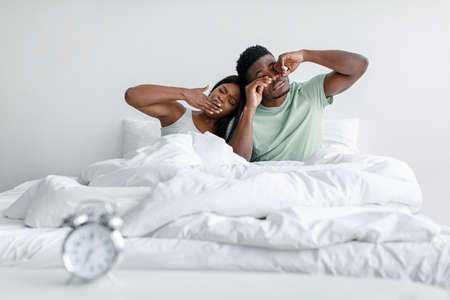 Sleepy millennial african american female and man yawns and rubs eyes sitting on soft comfort bed in...