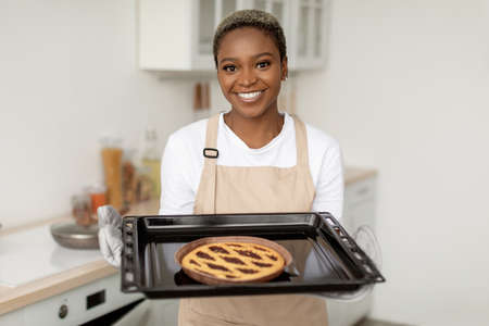 Glad smiling millennial african american woman in apron showing fresh tasty pie in minimalist kitchen interiorの写真素材