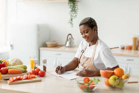 Cheerful millennial black lady in apron makes notes on table with organic vegetables and fruitsの写真素材