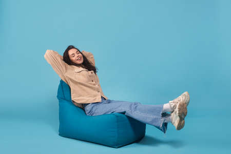 Satisfied happy millennial european woman in casual relaxes and enjoys rest on bagchair isolated on blue backgroundの写真素材