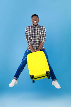 Time to travel. Full length of excited young black man jumping with bright suitcase over blue studio backgroundの写真素材
