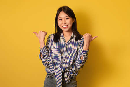 Joyful young Asian woman in casual wear showing thumbs up with both hands, smiling at camera on...