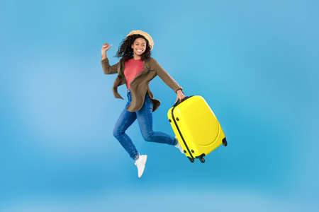 Excited young black woman in casual wear with bright suitcase jumping on blue studio background, copy spaceの写真素材