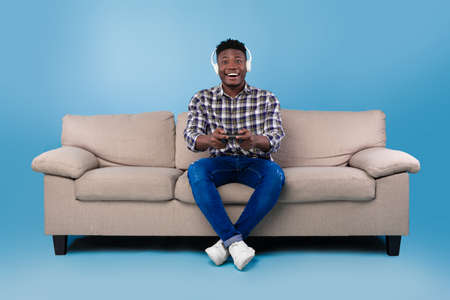 Happy young black guy with joystick playing video game, wearing headset, sitting on couch over blue...