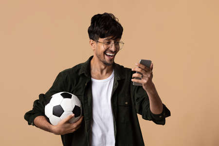 Excited indian guy with soccer ball using mobile phone