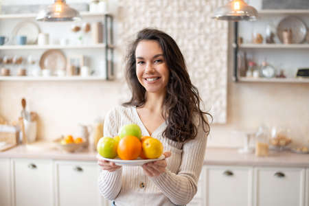 Happy woman holding plate with fresh fruits, looking and smiling at camera, standing in kitchen interiorの写真素材