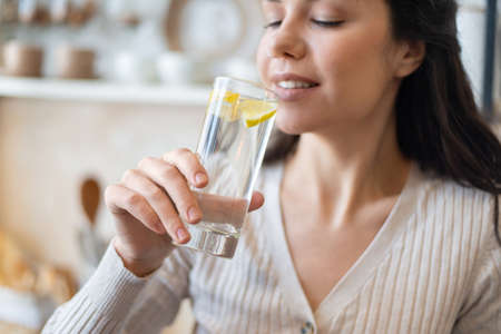 Refreshment concept. Young woman drinking water with lemon, holding glass, standing in kitchen,...