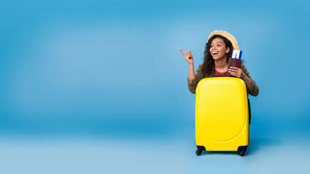Carefree young black woman holding passport and tickets, sitting behind suitcase, ready to travel on blue backgroundの写真素材