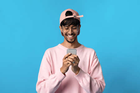 Cheerful hindu guy hipster using mobile phone on blueの写真素材