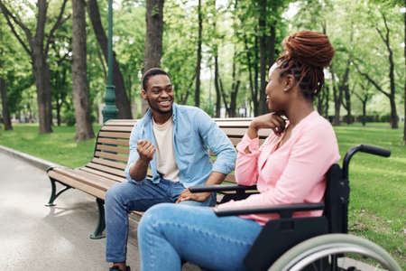 Cheery young black woman in wheelchair speaking to her boyfriend at urban park, copy spaceの写真素材