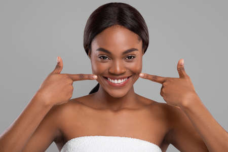 Cheerful pretty black woman wrapped in towel pointing fingers at perfect snow white smile