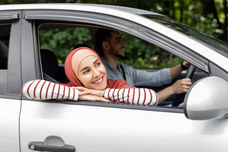 Smiling millennial arab female in hijab ride in car looks out the open window outdoor, enjoy journeyの写真素材