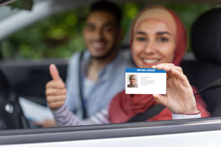 Glad middle eastern male and female in hijab show driving license and thumb up in open window in car after examの写真素材