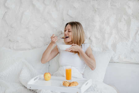 Happy pretty hungry young european blonde woman wake up, have breakfast in white bedの写真素材