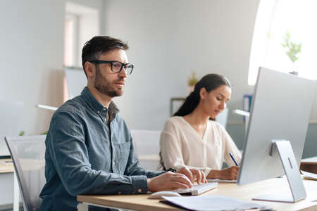 Focused Caucasian man using computer, working in modern office with his colleague, copy spaceの写真素材