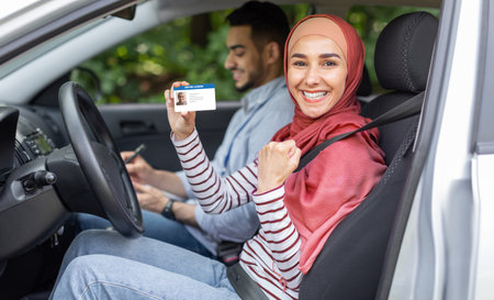 Glad muslim woman in hijab show driving license and fist in open door in car after exam with male...