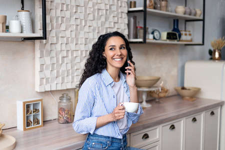 Happy latin woman drinking coffee and having phone call, standing in kitchen interior and smilingの写真素材