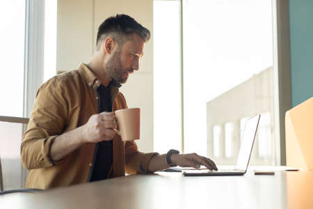 Man Working On Laptop Drinking Coffee In Office, Side View