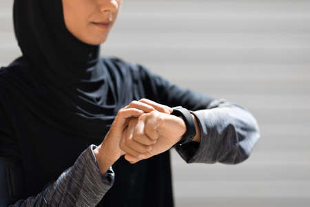 Confident young arab lady athlete in hijab checks pulse on fitness tracker on wall background,...
