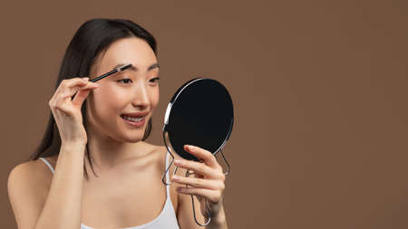 Natural makeup concept. Happy asian lady brushing eyebrows, holding brush and mirror, brown...