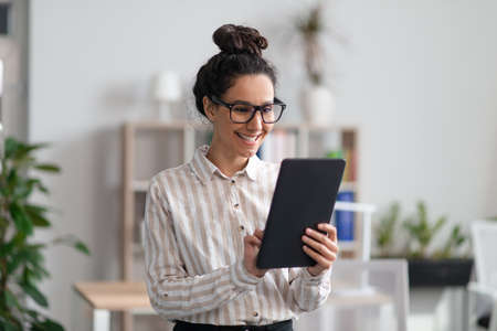 Positive young female entrepreneur using digital tablet in office and smiling, checking her agenda...
