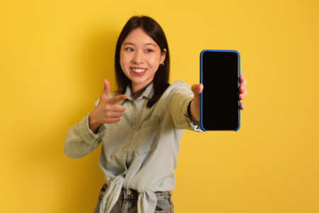 Cheery young Asian woman pointing at cellphone with empty space for mobile app or website on screen,...