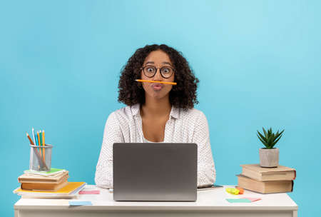 Silly young black female student sitting at desk with books and laptop, playing game with pencil on...