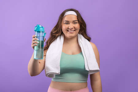 Happy young european plus size woman in sportswear with towel showing bottle of water