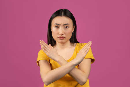 Portrait of serious asian lady showing stop gesture with crossed hands, posing over violet studio backgroundの写真素材