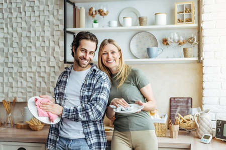 Cheerful young european lady and male washing dishes in modern kitchen interior. Hygiene, household chores in spare timeの写真素材