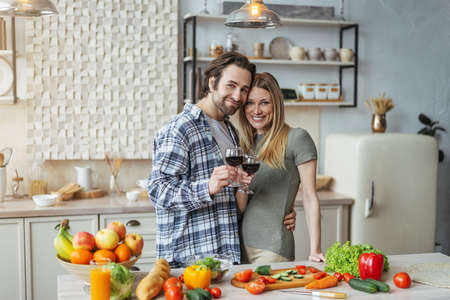 Cheerful young european husband with stubble hugs wife, clink glasses of wine, prepare food